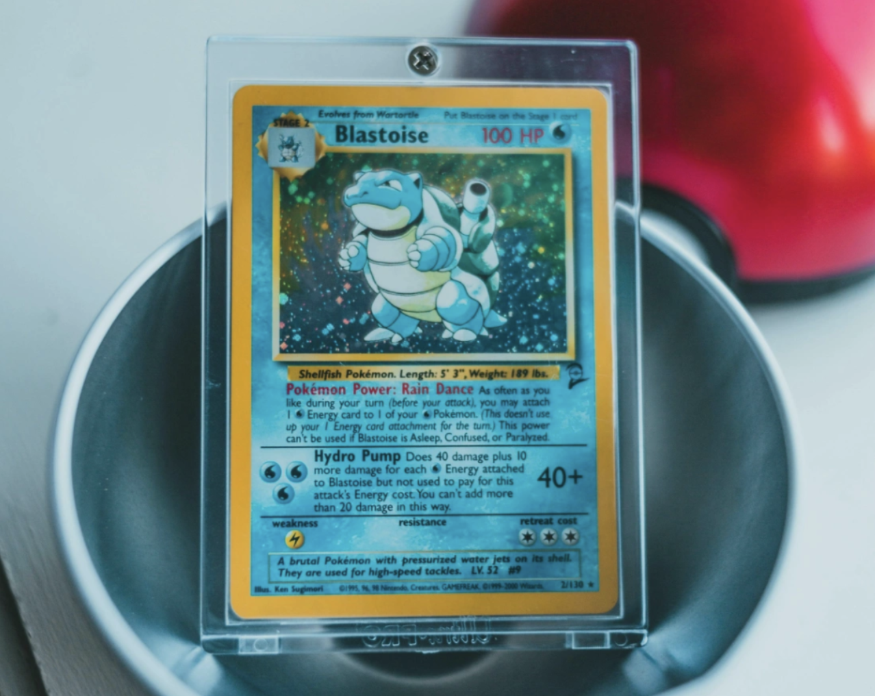 A holographic Blastoise Pokémon card in a plastic protective case, displayed upright in a metal bowl.